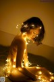 A naked woman sitting on a bed covered in lights.